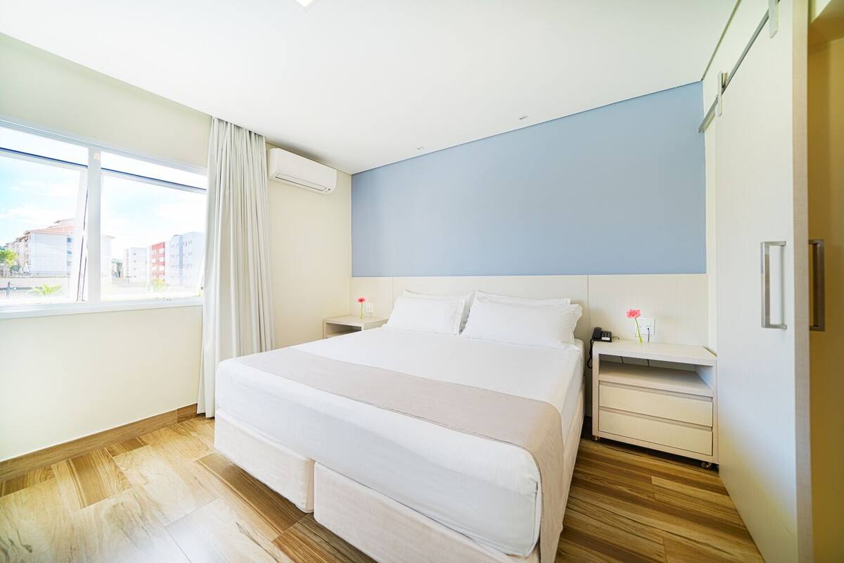 Una habitación de hotel moderna con una cama grande, una mesita de noche y un equipo de aire acondicionado, con vista a la ciudad a través de la ventana.