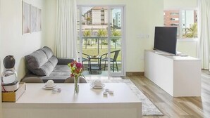 Un salón moderno con sofá, una mesa de centro con un florero, televisor en un mueble bajo y un balcón con vistas a edificios.