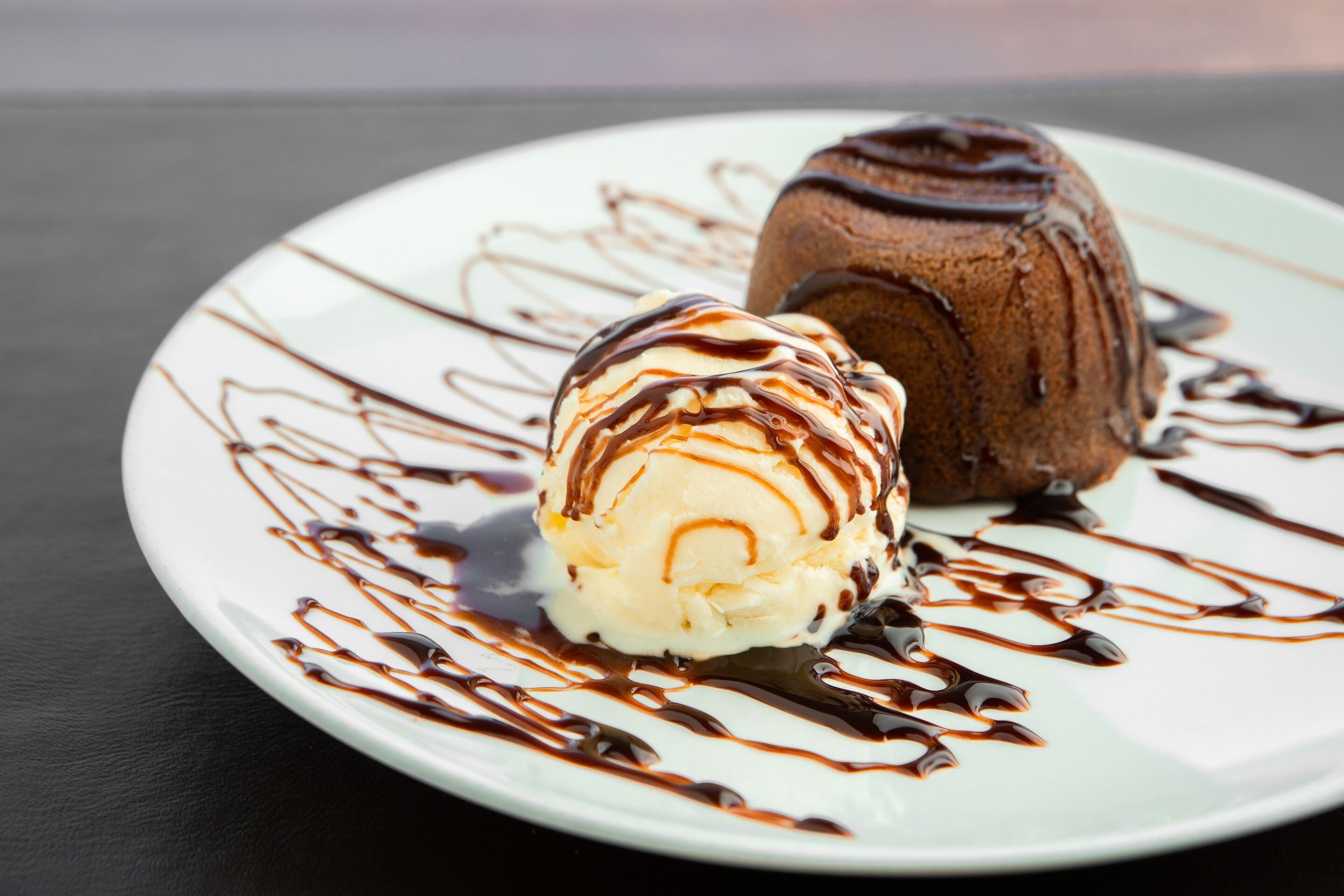 Un plato de postre con una bola de helado de vainilla y un postre de chocolate, rociado con salsa de chocolate.