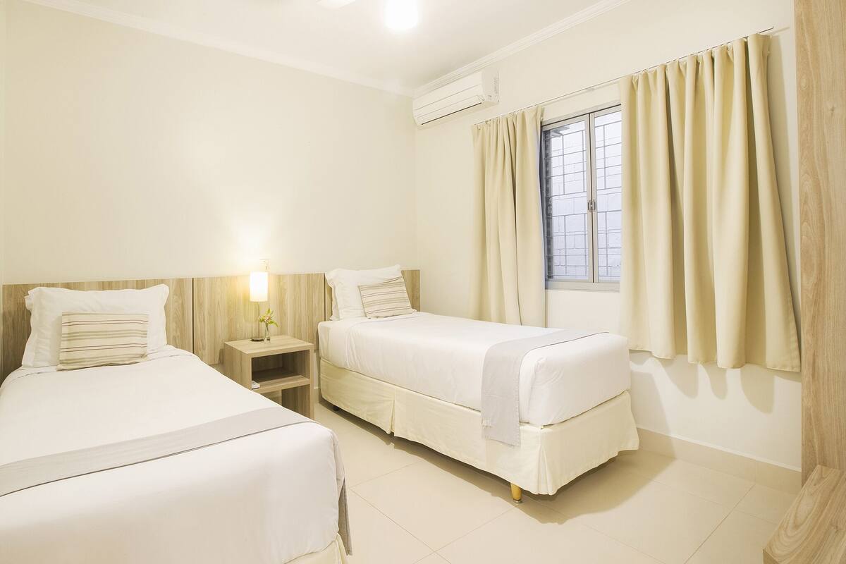 Una habitación de hotel con dos camas, una mesita de noche con lámpara, una ventana con cortinas y un ventilador de techo.
