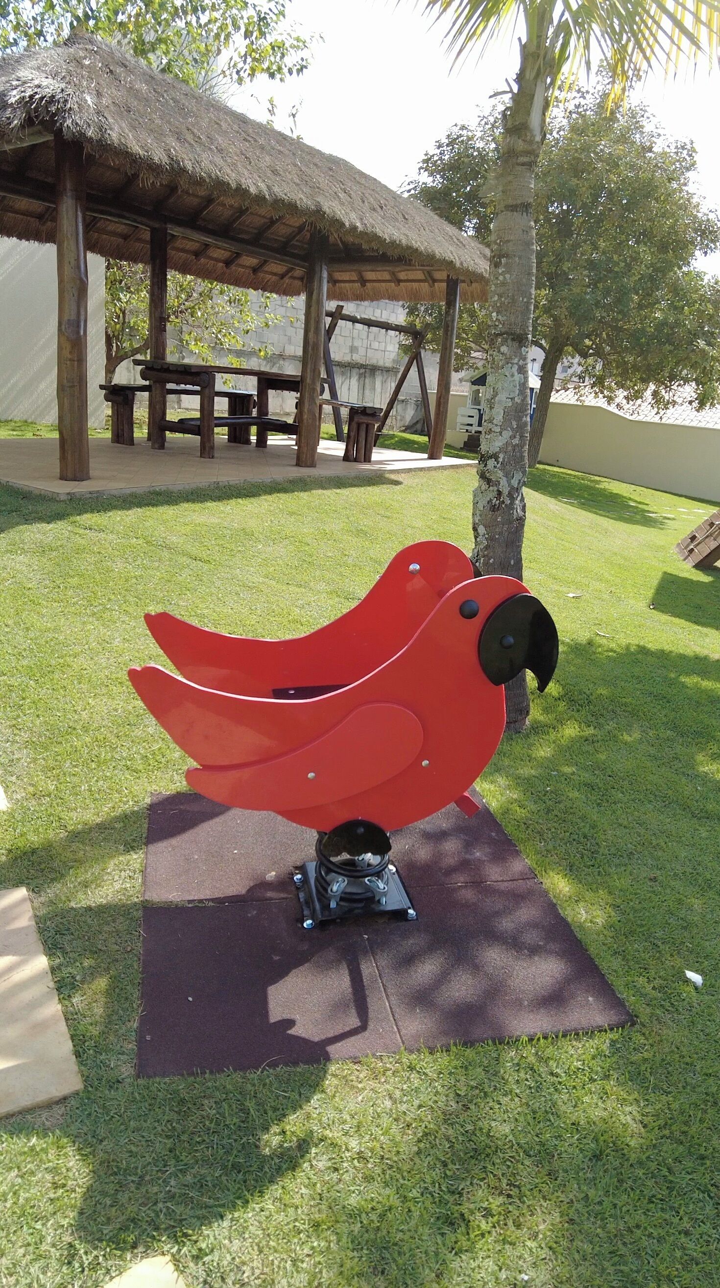 Een rode, vogelvormige sculptuur op een vierkant platform in een park, met een rieten paviljoen op de achtergrond.