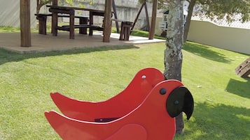 A red bird-shaped sculpture on a square platform in a park with a thatched-roof pavilion in the background.