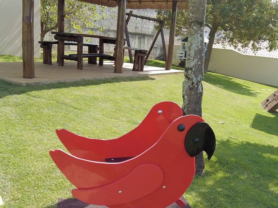 A red bird-shaped sculpture on a square platform in a park with a thatched-roof pavilion in the background.