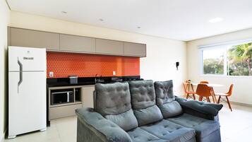 A modern kitchen with a refrigerator, oven, and microwave. There is a gray sofa and orange chairs in the living area.