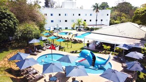 Um hotel com várias piscinas, espreguiçadeiras, guarda-sóis e um edifício grande ao fundo.