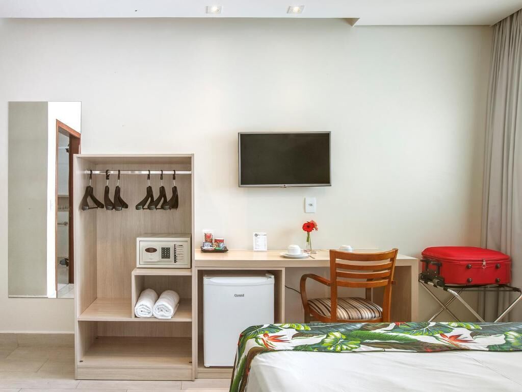 A hotel room with a bed, a desk, a chair, a TV, a wardrobe, and a red suitcase.