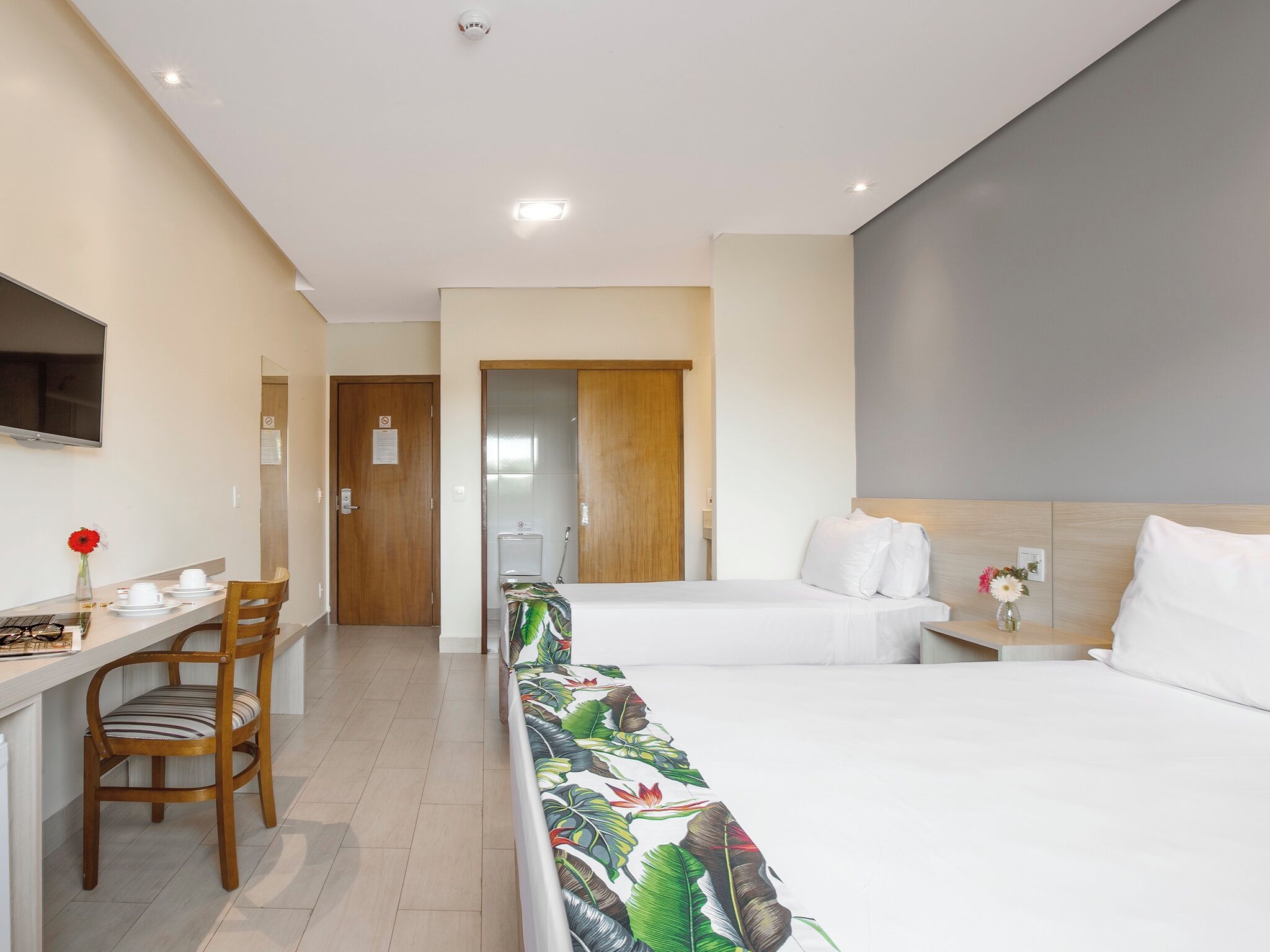 A hotel room with a bed, a desk with a chair, a TV, and a bathroom visible through an open door.