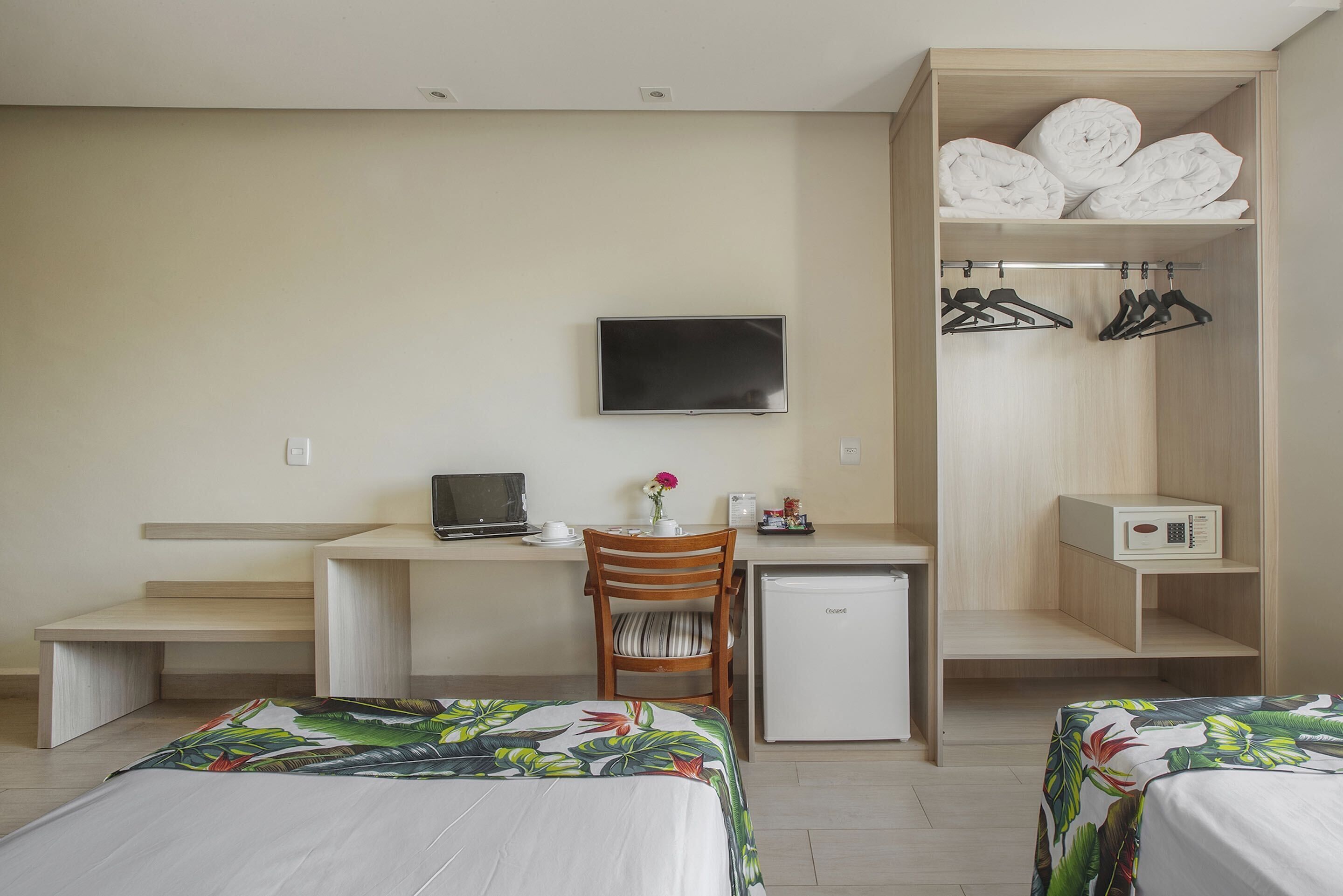 Habitació d'hotel amb llit, escriptori amb ordinador, televisió, nevera petita, cadira i armari amb penjadors.