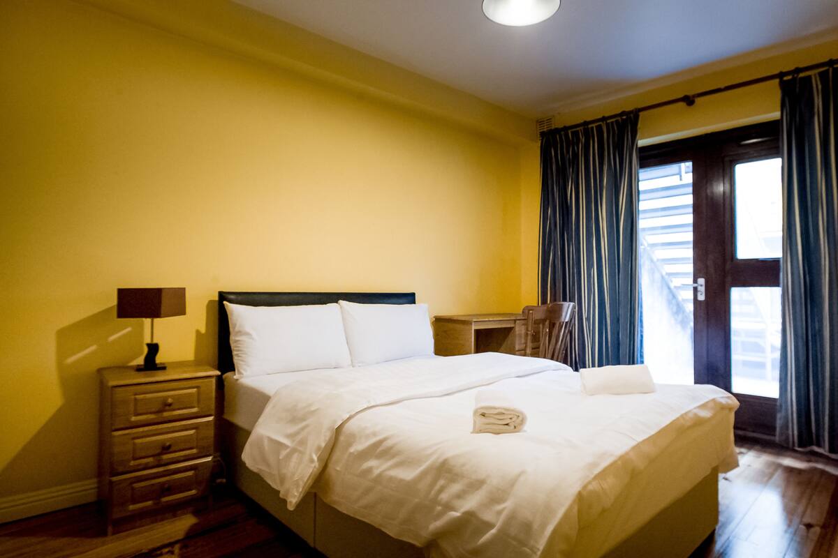 A hotel room with a bed, a nightstand, a lamp, and a view of the outside through a door.