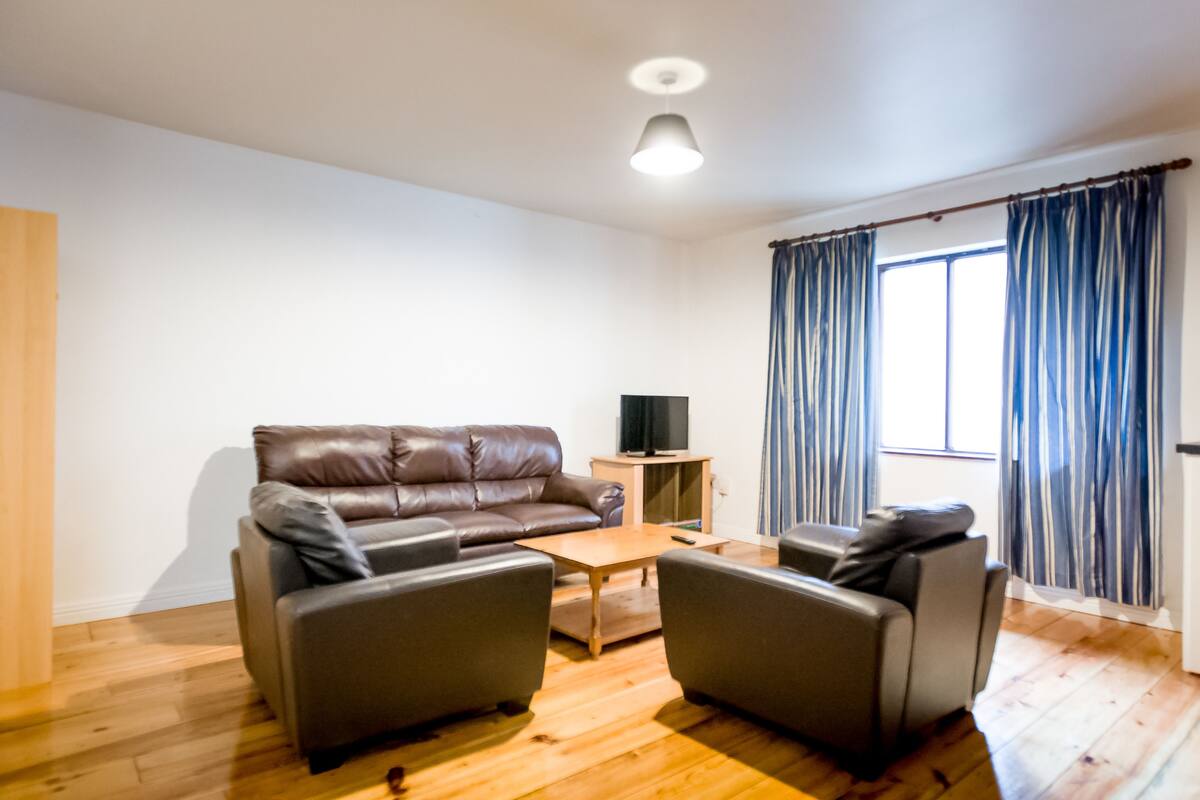 A modern living room with a leather sofa, two armchairs, a wooden coffee table, and a television on a stand.