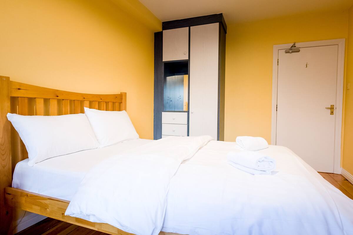 A neatly made bed with white linens and a wooden headboard in a room with yellow walls and a white door.