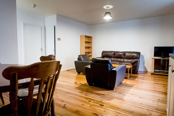 A modern living room with a wooden floor, a brown leather sofa, a grey armchair, a wooden dining table with chairs, and a television on a stand. - West end Apartments Galway (Galway)