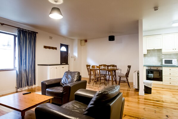 A living room with a wooden coffee table, two leather sofas, a dining table with chairs, a kitchen area with cabinets and a microwave, and a window with curtains.