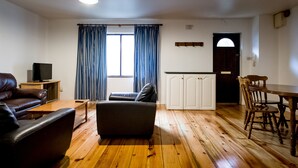 Ein Wohnzimmer mit Sofa, Couchtisch, Fernseher auf einem Schrank, Esstisch mit Stühlen und einem Fenster mit Vorhängen.