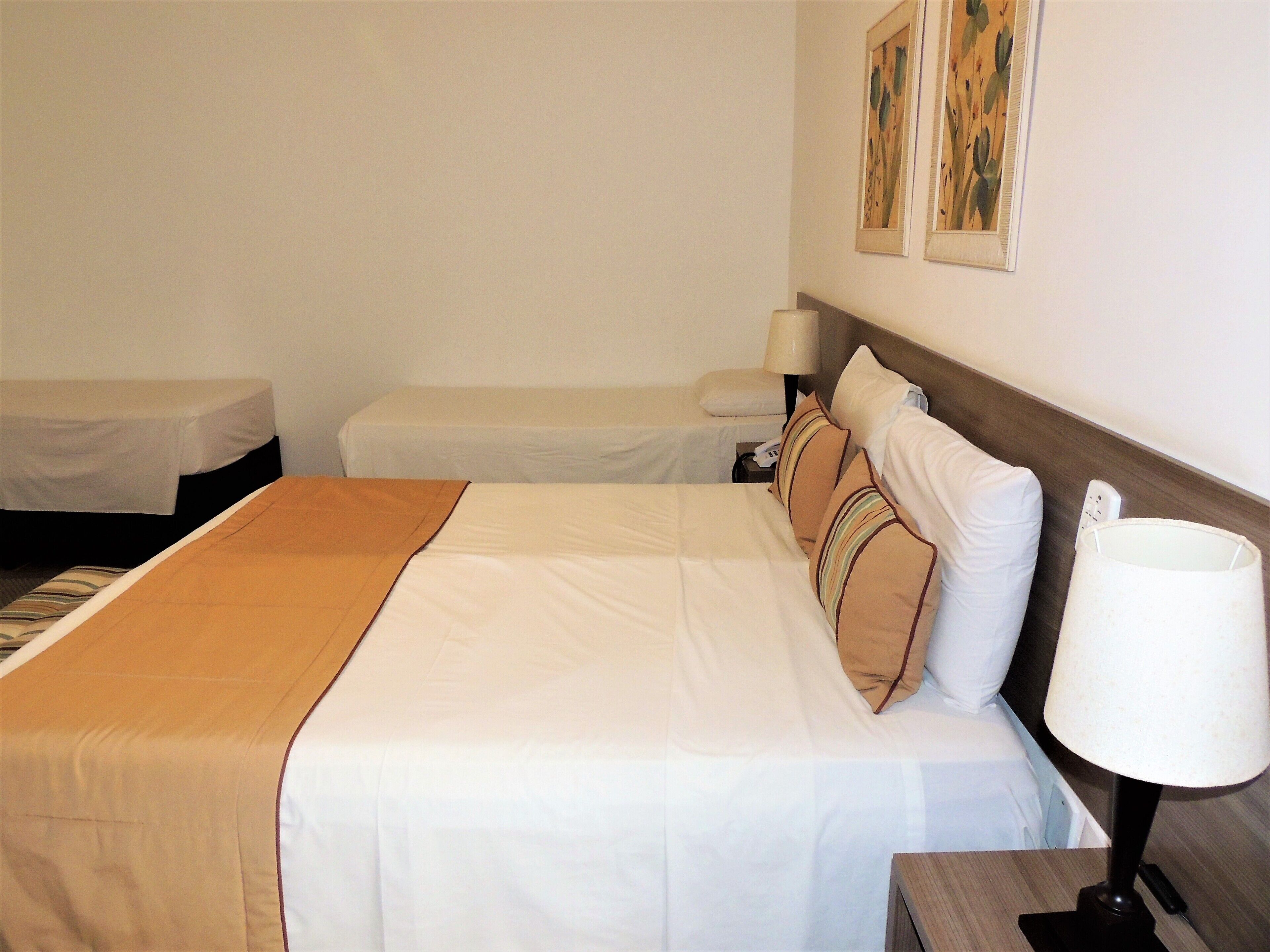 Ein Hotelzimmer mit einem großen Bett, zwei Einzelbetten, einem Nachttisch mit Lampe und einem Kunstwerk an der Wand.