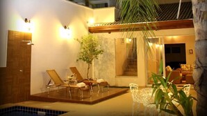 Un área de piscina moderna al aire libre con sillones de descanso y una mesita pequeña, rodeada de paredes blancas y una terraza de madera.