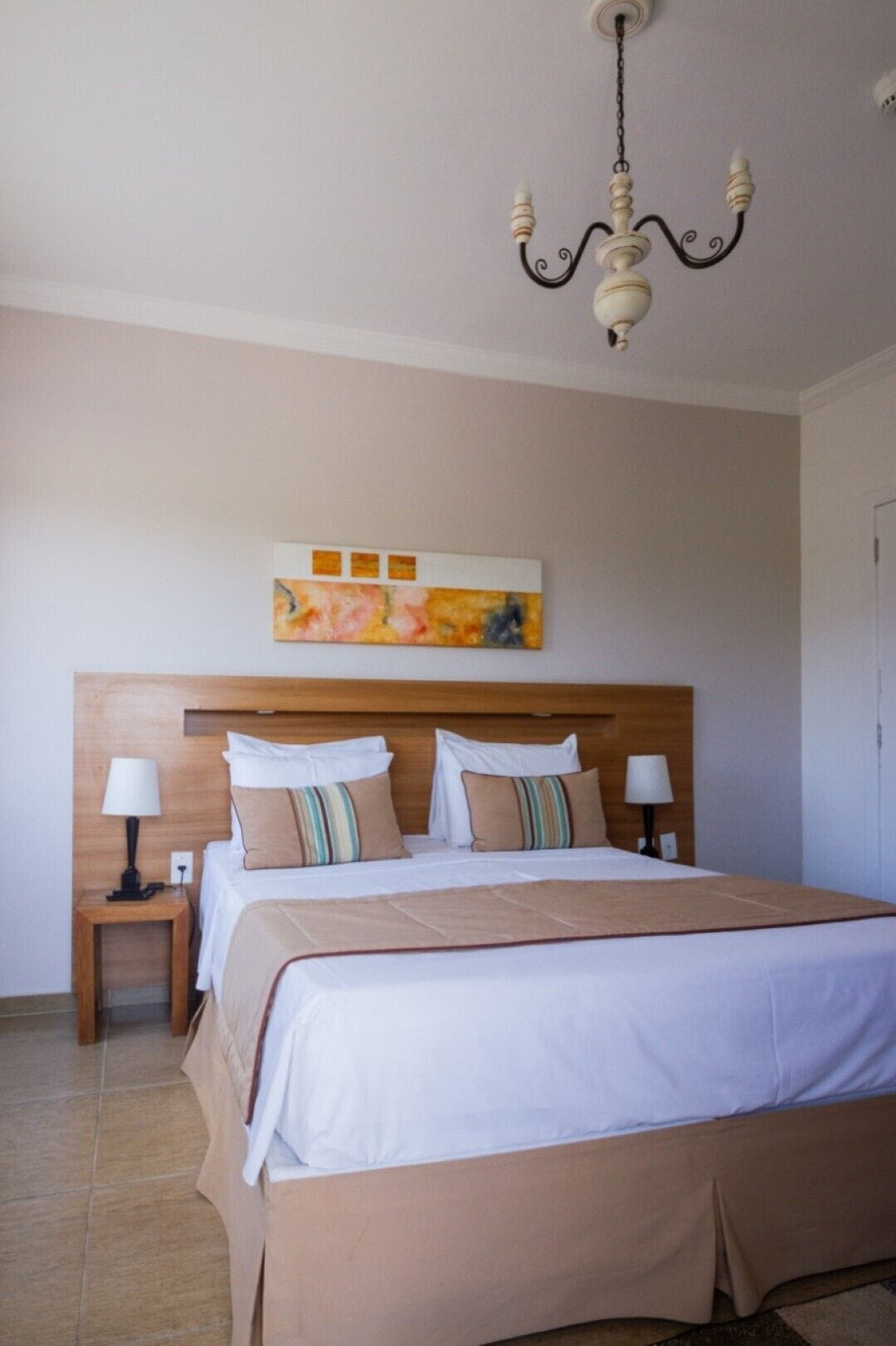 A neatly arranged hotel room with a large bed, two bedside tables, a chandelier, and a wall-mounted artwork.