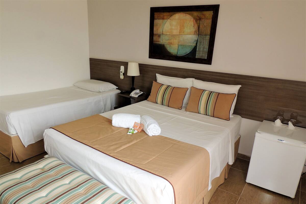 A hotel room with two single beds, a double bed, a bedside table with a lamp, a small refrigerator, and a painting on the wall.