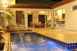 Una piscina con fuente de agua, un patio cubierto con mobiliario exterior y vista a una sala de estar.