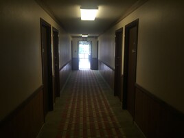 Un pasillo largo y estrecho con alfombra estampada, puertas de madera a ambos lados y una señal de salida brillante al final.