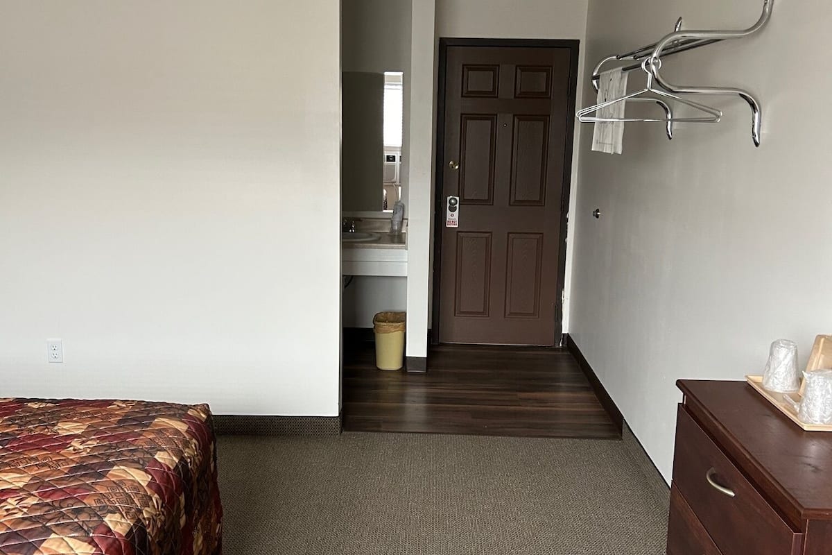 A hotel room with a bed, a dresser, a coat rack, and a bathroom visible through an open door.