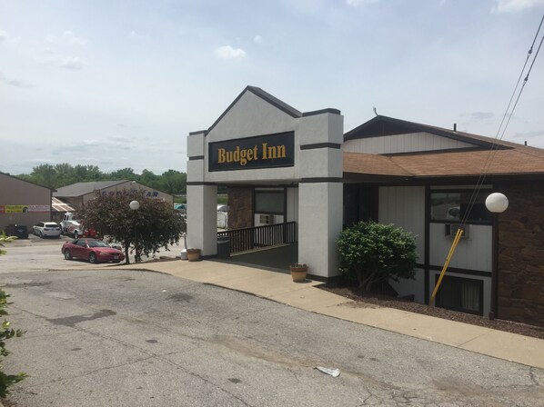 A Budget Inn with a simple, modern design, featuring a prominent sign, a parking lot with several cars, and a small landscaped area with a tree and potted plants.