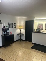 Un vestíbulo de hotel con mostrador de recepción, una máquina de café, una pequeña mesa con lámpara y obras de arte enmarcadas en las paredes.
