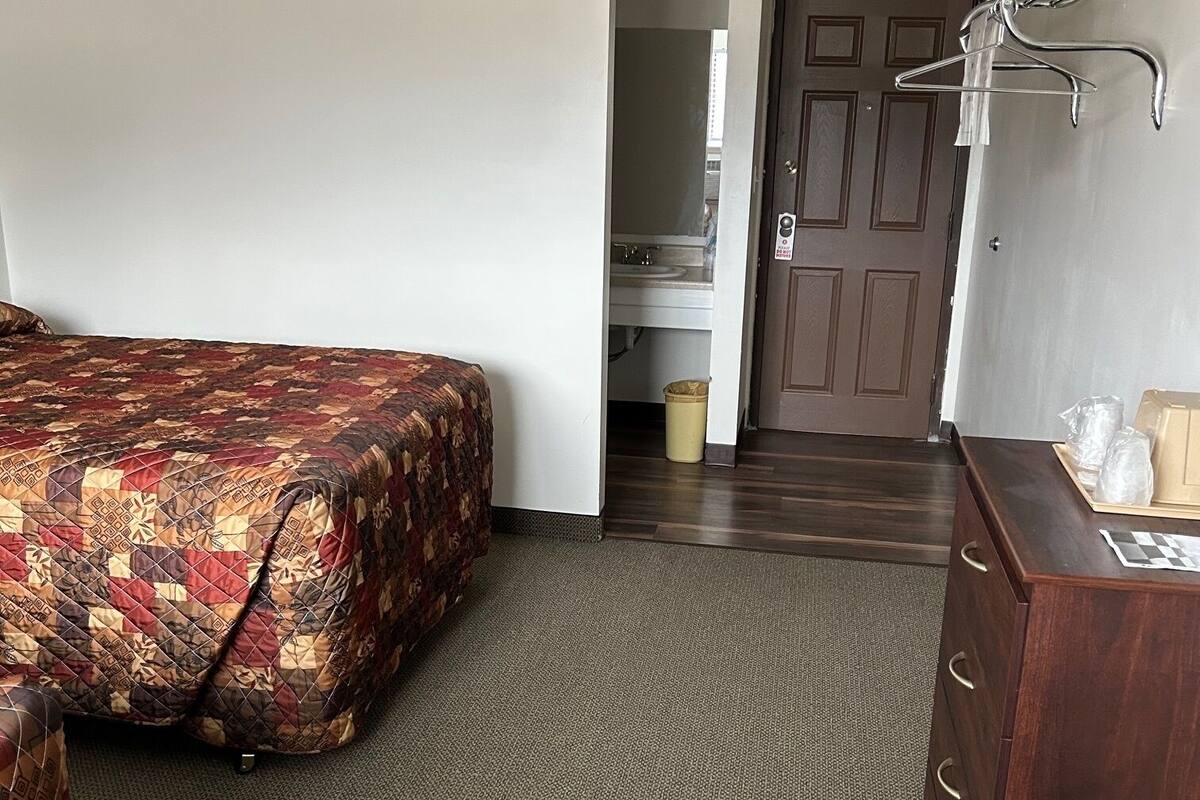 A hotel room with a bed, a dresser, a trash can, and a door leading to a bathroom.