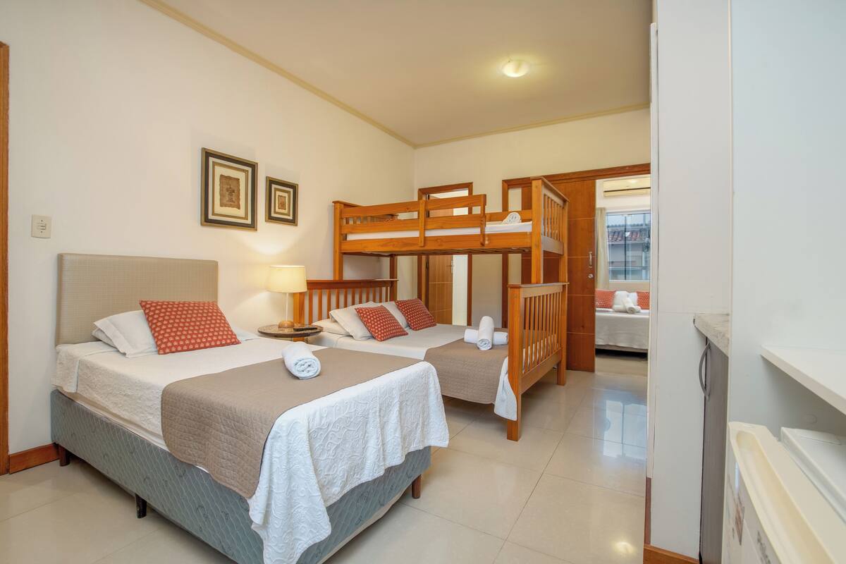 A hotel room with two beds, a wooden bunk bed, a bedside table with a lamp, and a view of a balcony.