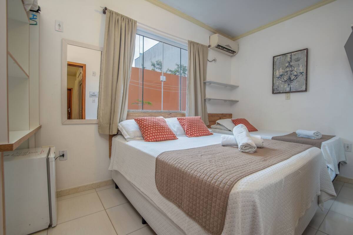 A hotel room with a large bed, a smaller bed, a window with curtains, a wall-mounted air conditioner, and a small refrigerator.