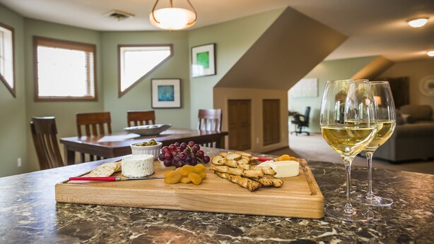 Un comedor con una mesa de mármol, una tabla de madera con queso, uvas y galletas, y una copa de vino blanco.