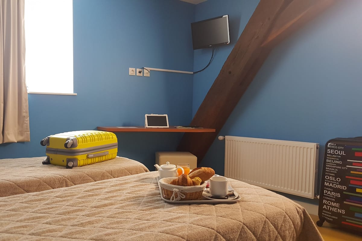 A room with two beds, a suitcase, a table with a teapot, a cup, and a basket of bread.