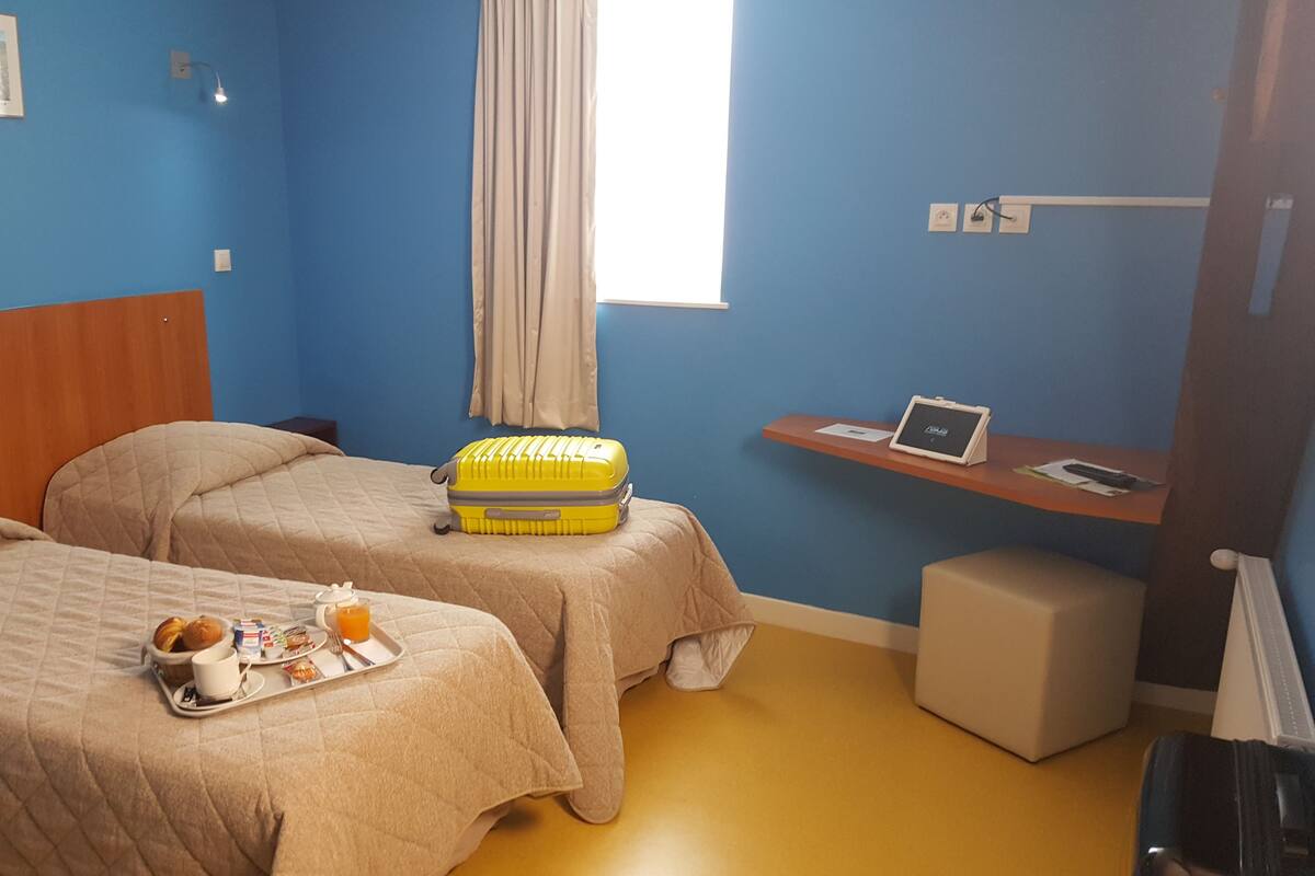 A hotel room with two beds, a small desk with a laptop, a window with curtains, and a tray with breakfast items.