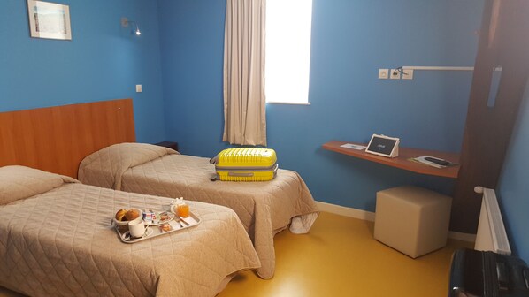 A hotel room with two beds, a small desk with a laptop, a window with curtains, and a tray with breakfast items.