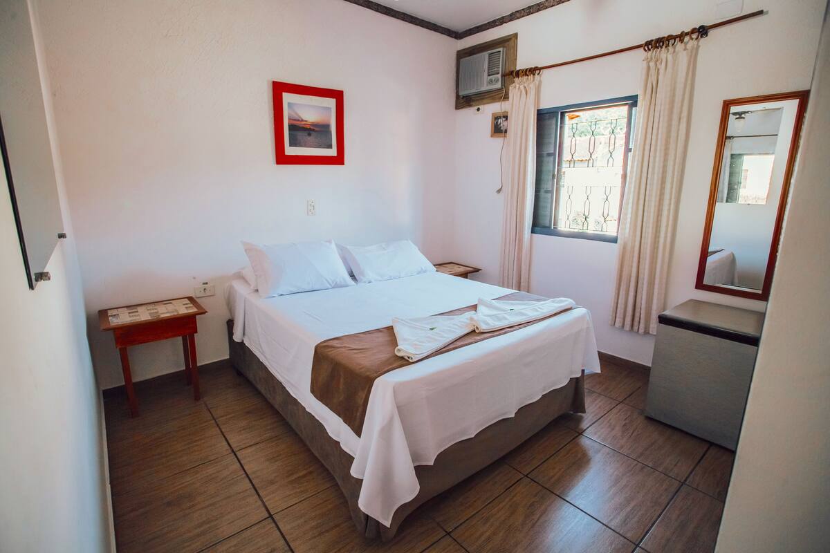 A bedroom with a bed, a nightstand, a small table, a window with curtains, a mirror, and a wall-mounted air conditioner.