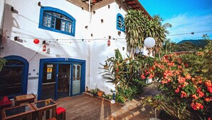 A white building with blue accents, wooden furniture, and a garden with red flowers. - Pousada Rafimar (Ilhabela)