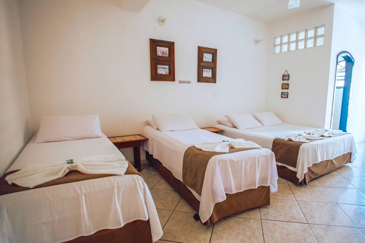 A hotel room with three single beds, each with a white bedspread and a brown blanket. There are framed pictures on the wall, a small clock, and a window with a view of the outside.