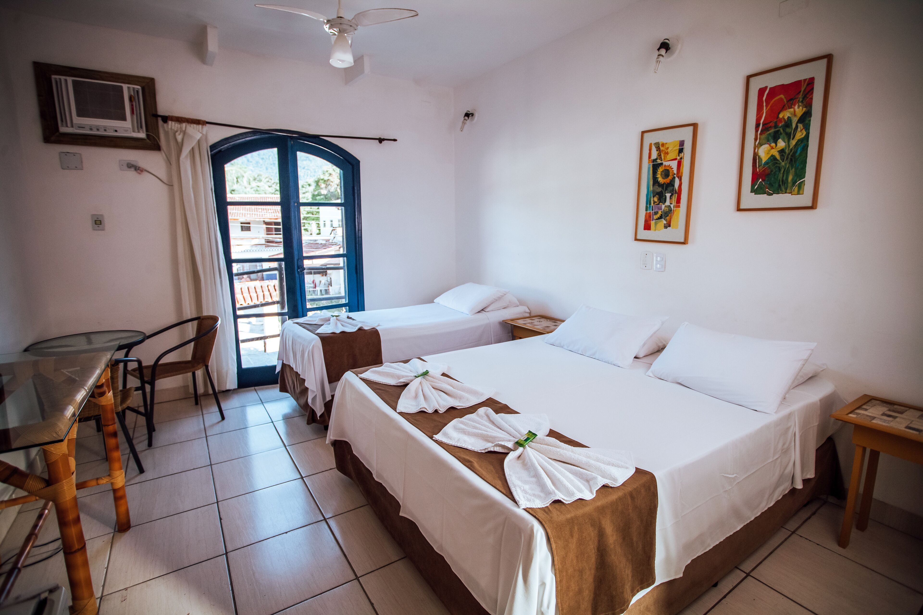 A hotel room with two beds, a small table, a chair, a ceiling fan, and a window with a view of buildings outside.