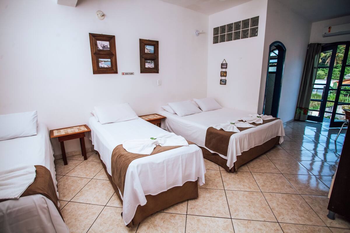 A hotel room with three beds, a tiled floor, and a view of greenery through the window.