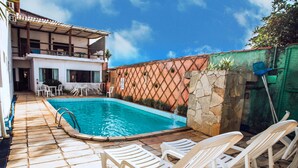 A swimming pool with white lounge chairs, a stone wall, and a building with a tiled roof in the background. - Pousada Rafimar (Ilhabela)