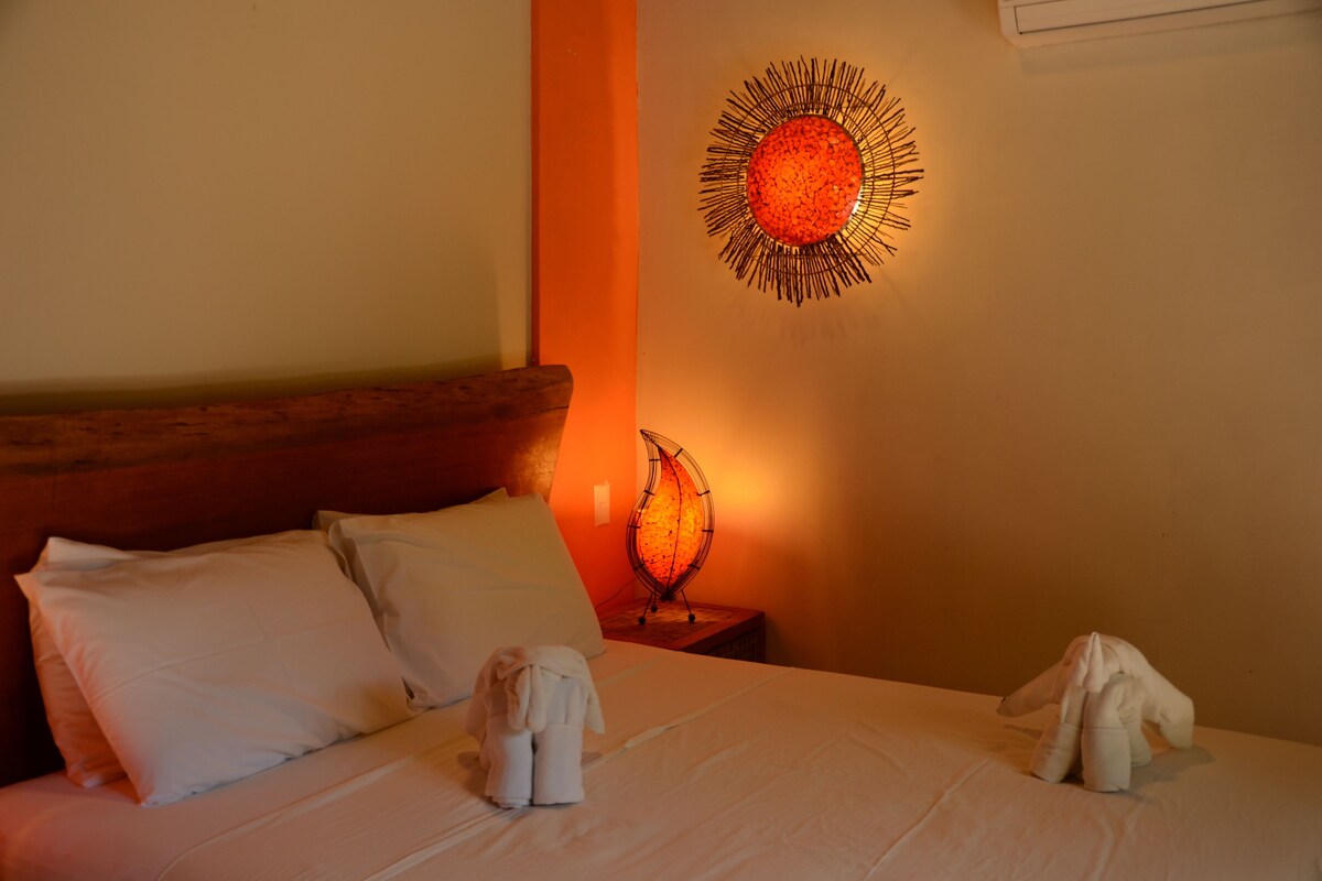 Een net opgemaakt bed met witte lakens en kussens, een decoratief kunstwerk aan de muur en een klein tafeltje met een lamp.