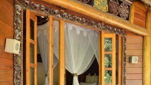 Een houten hut met rieten dak, een bed met hemelbed en een decoratief houten paneel boven de ingang.