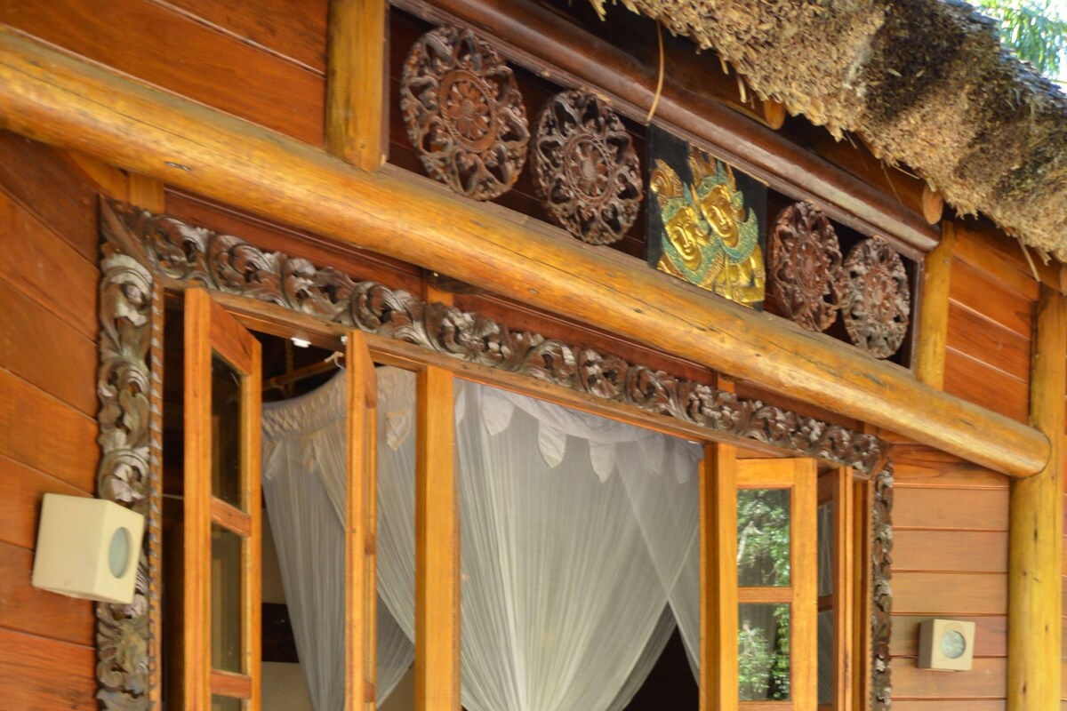 Een houten hut met rieten dak, een bed met hemelbed en een decoratief houten paneel boven de ingang.