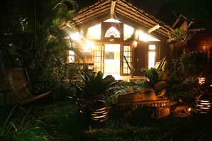 Een goed verlicht houten huis met een veranda, omringd door weelderige begroeiing en decoratieve verlichting.