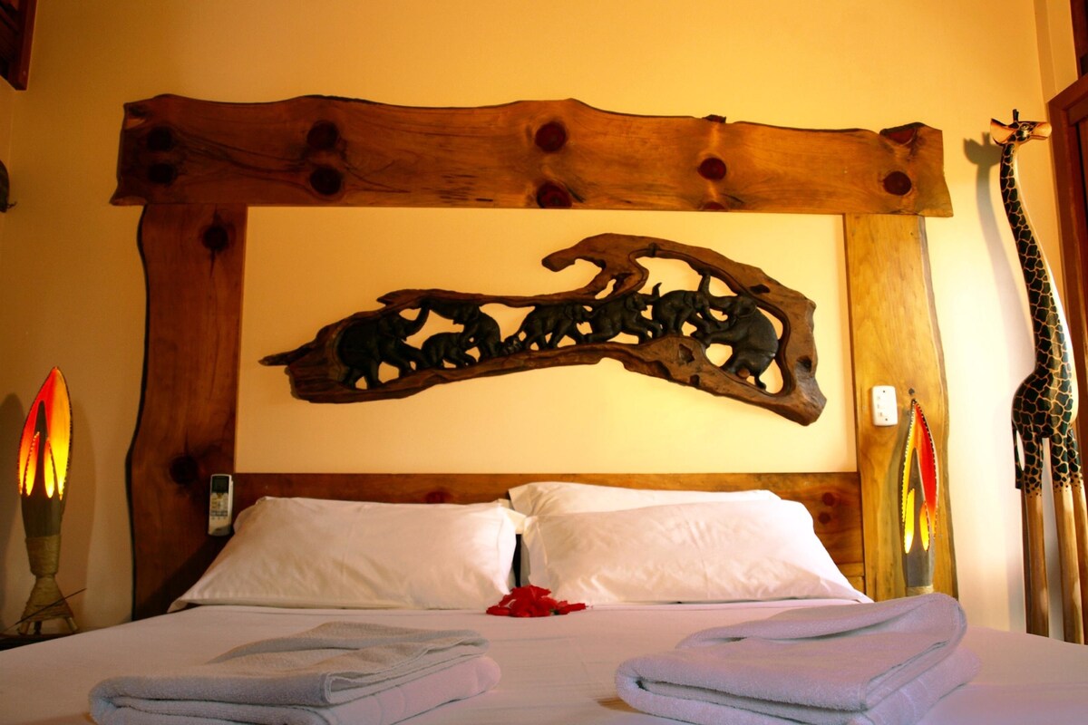 Een net opgemaakt bed met opgevouwen handdoeken, een houten hoofdbord met een gedecoreerd ontwerp en een kunstwerk aan de muur.