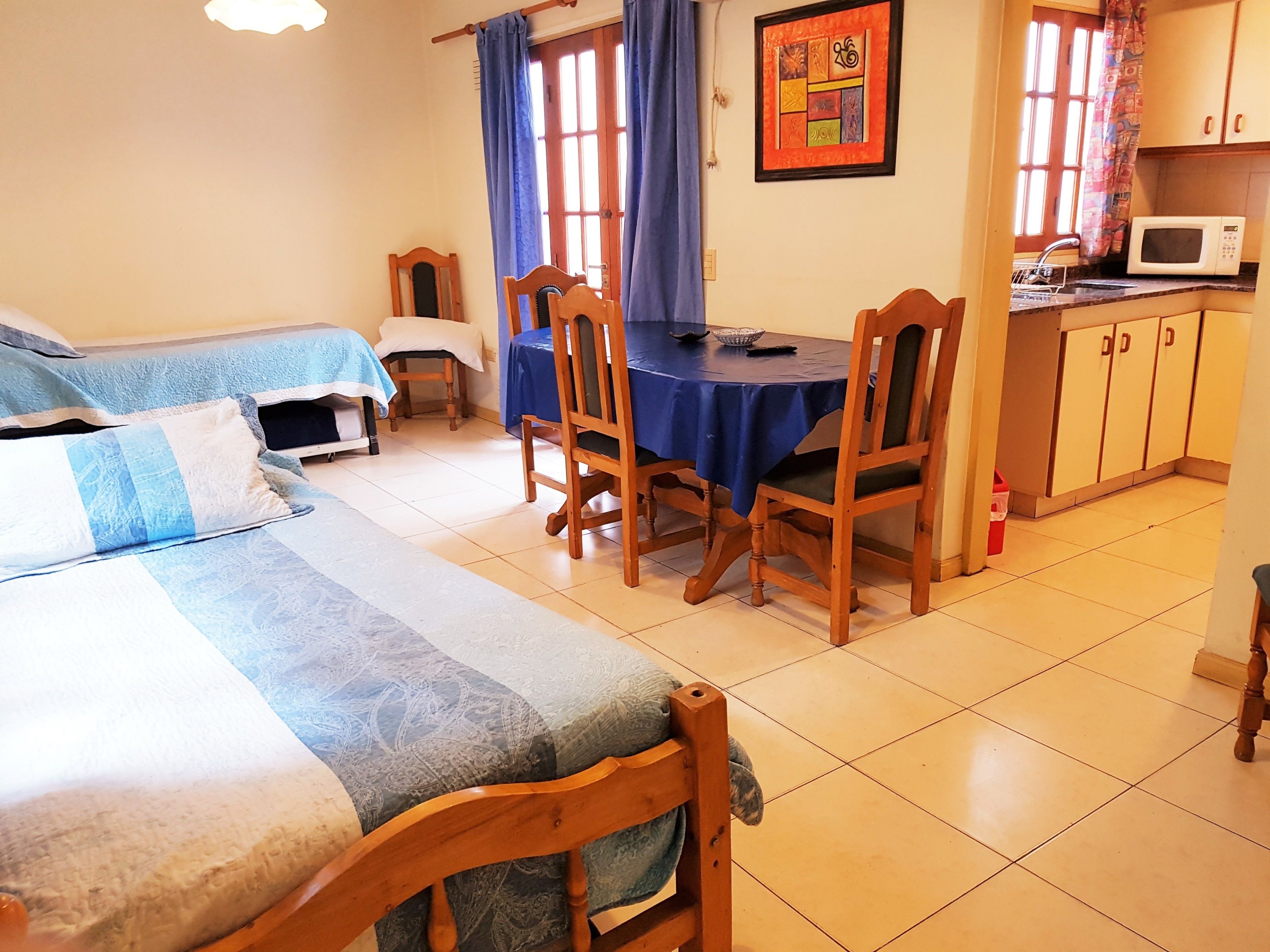A room with two beds, a dining table with chairs, a small kitchen area with a microwave, and a window with curtains.
