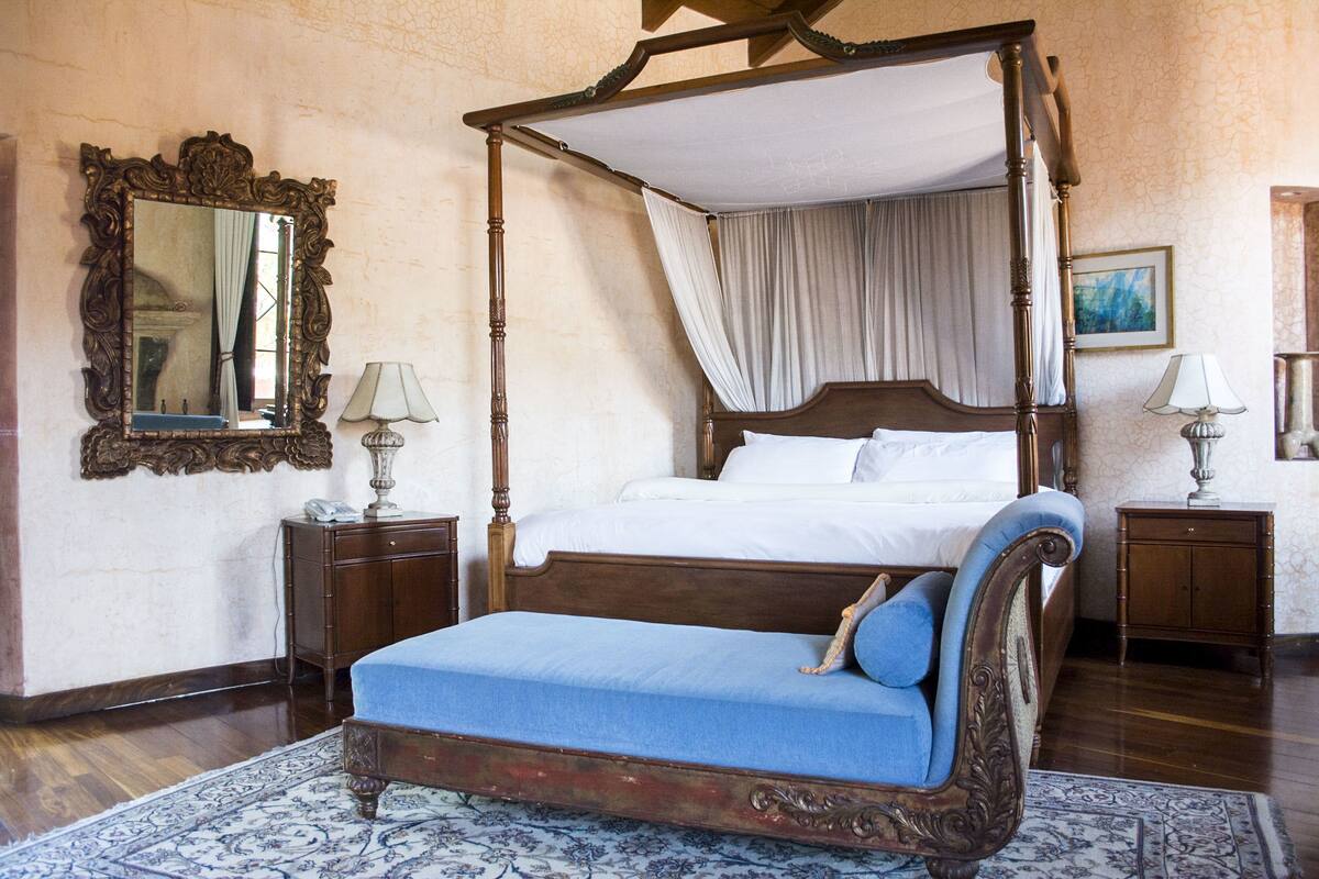 Una cama con dosel y cuatro columnas, con un banco tapizado en azul, mesitas de noche de madera con lámparas y un espejo grande y ornamentado en la pared.