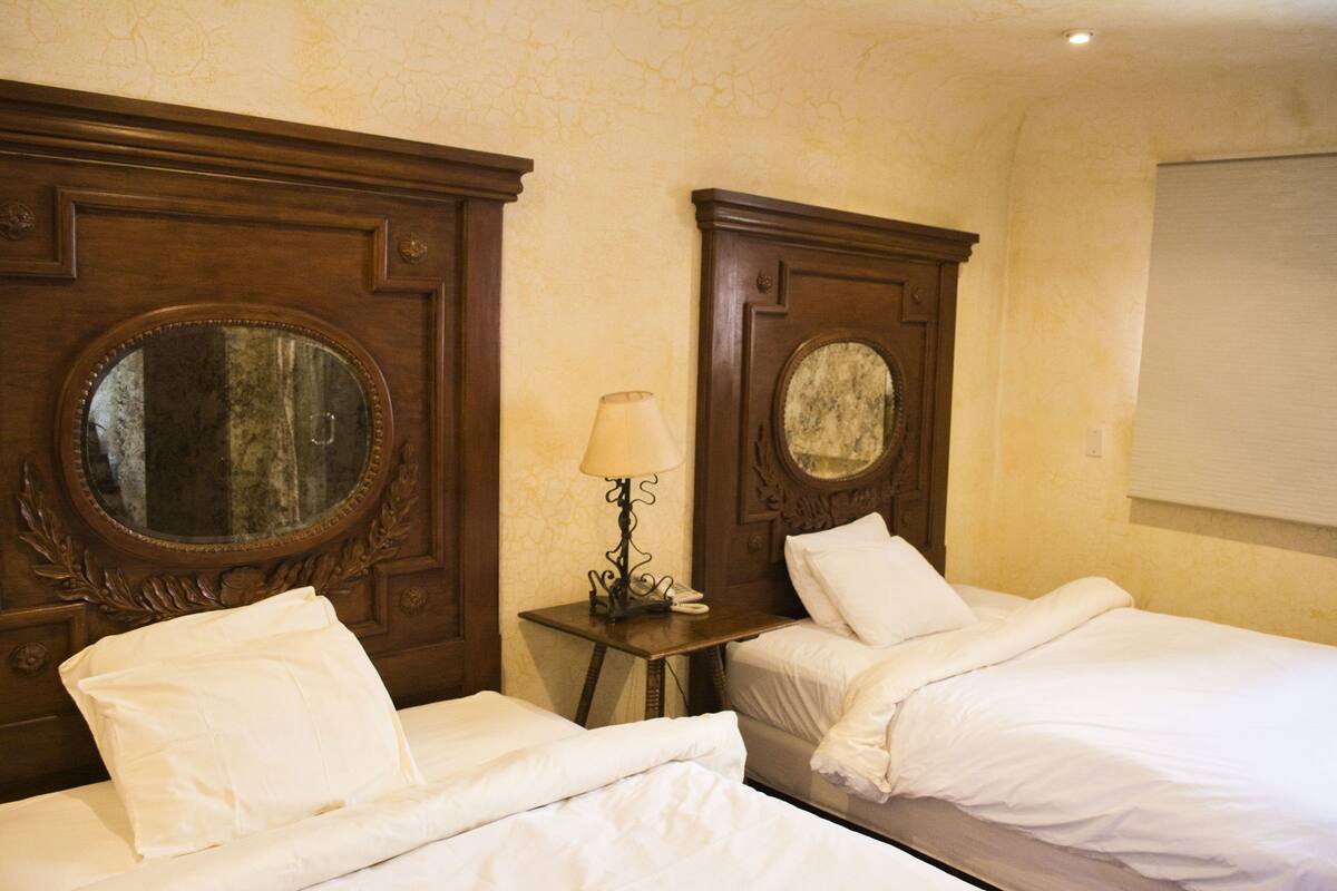 Habitación de hotel con dos camas, una mesita con lámpara y un espejo grande en la cabecera.