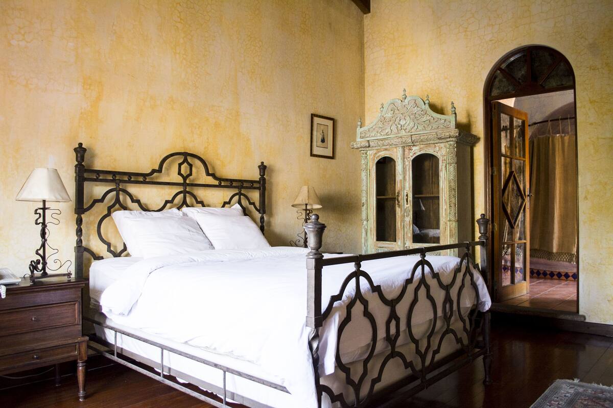 Un dormitorio con una cama con dosel, una mesita de noche con lámpara y un aparador decorativo.