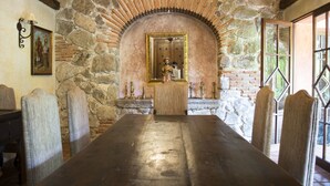 A long, dark wooden table in a stone-walled room with a mirror and religious statues. - Posada de Los Leones (Antigua Guatemala)